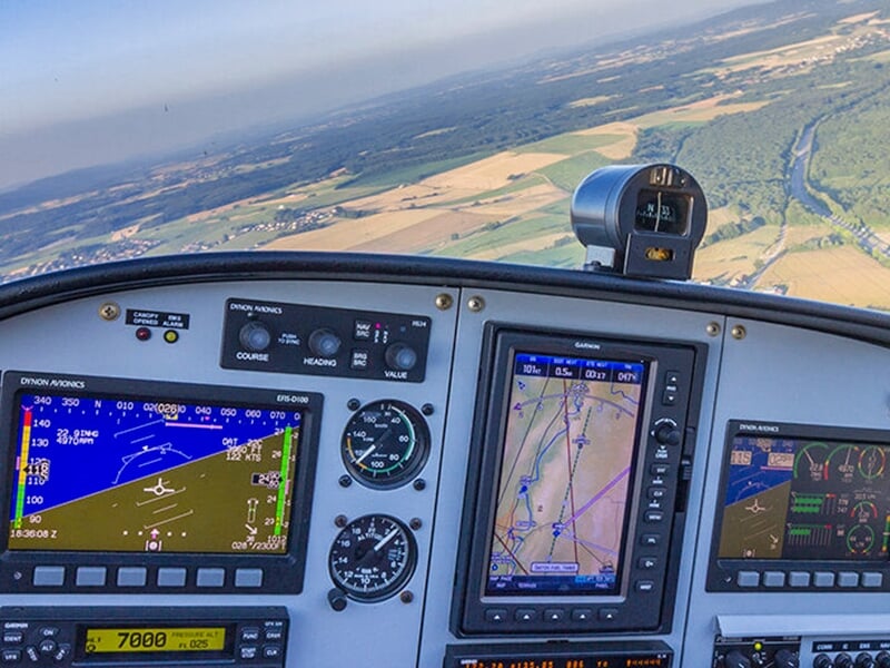 Tableau de bord d'un avion en vol avec écrans et instruments de navigation, offrant une vue sur la campagne en contrebas.