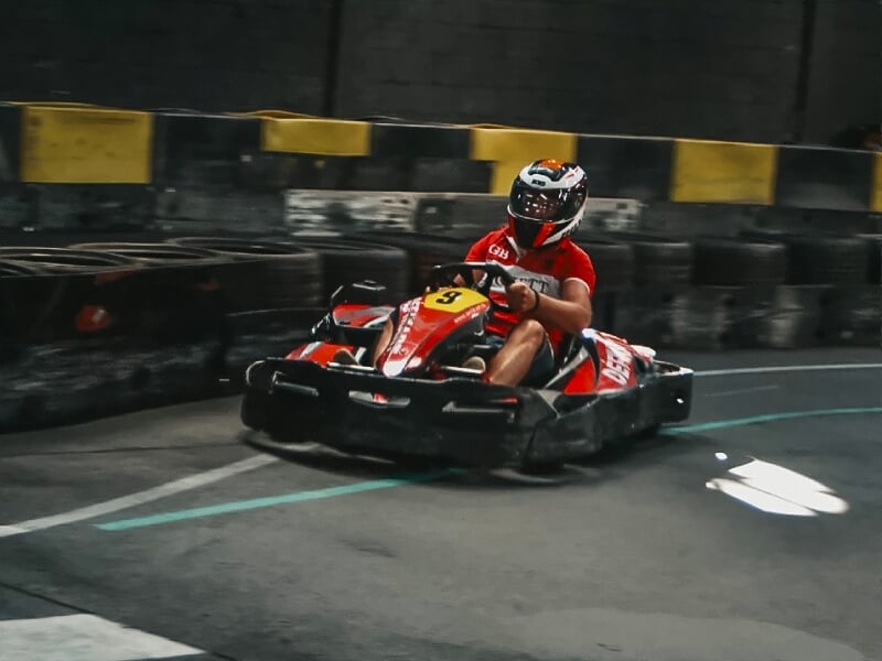 Un pilote en casque et tenue rouge conduit un karting rouge sur une piste de karting intérieure avec des barrières en pneus.