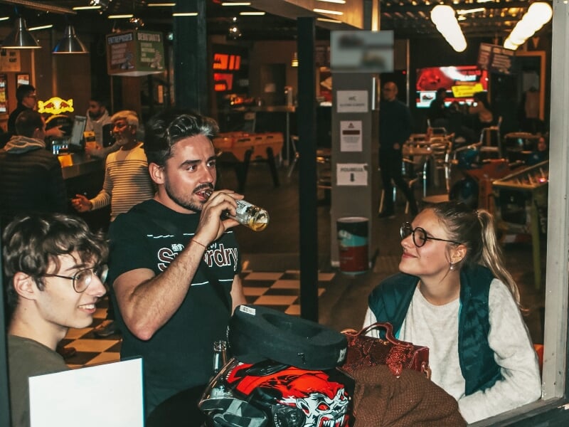 Trois personnes discutent dans un espace intérieur, une boit une boisson, avec un casque de karting posé sur la table.