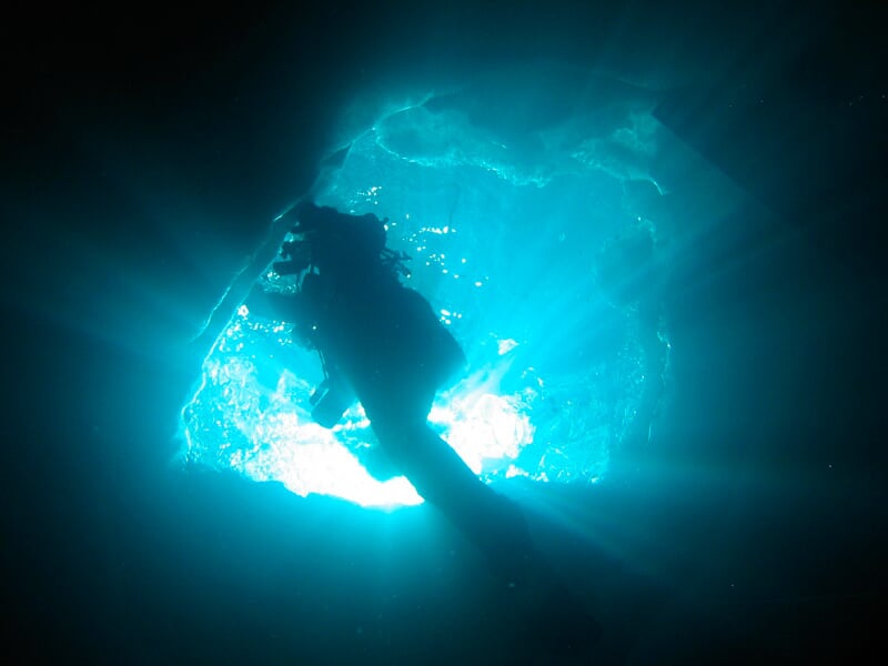 Silhouette d'un plongeur sous-marin nageant vers la lumière à travers une ouverture rocheuse.
