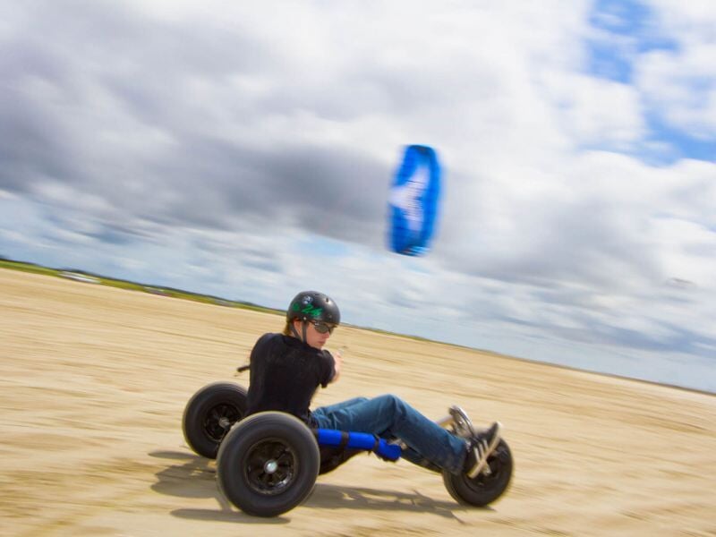 Personne en casque pilotant un buggy de kite sur une plage sablonneuse sous un ciel nuageux.