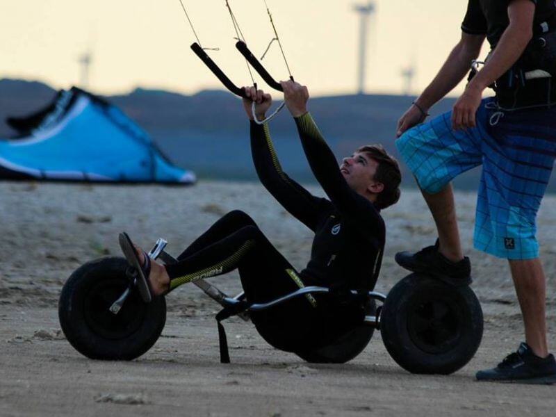 Jeune homme assis sur un buggy de kite, prêt à décoller avec une aile bleue sur la plage.
