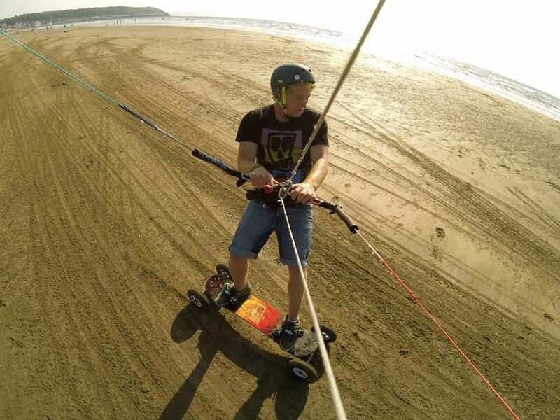 Homme en short et casque tenant une barre de kite, debout sur un mountainboard sur la plage.