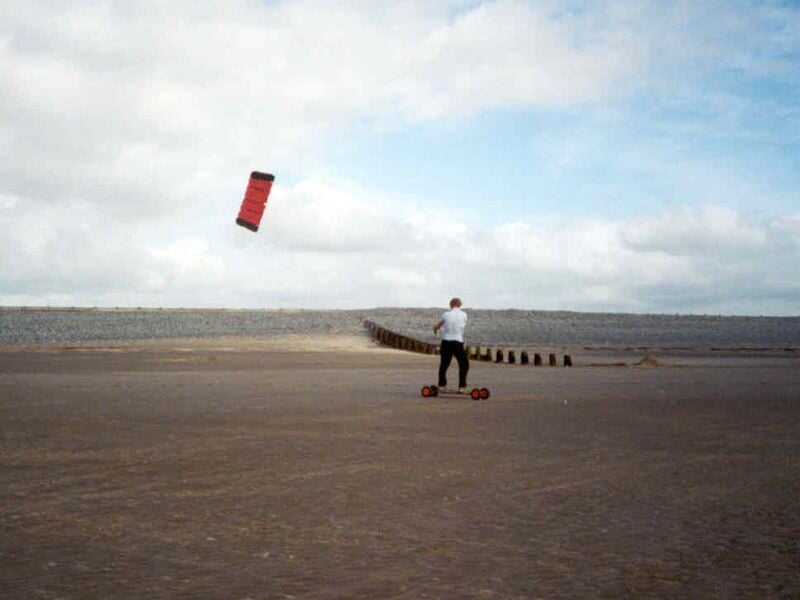 Personne sur un mountainboard avec une aile rouge, roulant sur une plage large et vide.