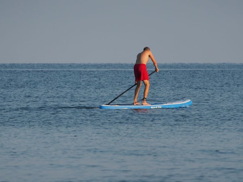Homme en short rouge pagayant debout sur un paddleboard sur une mer calme et bleue.
