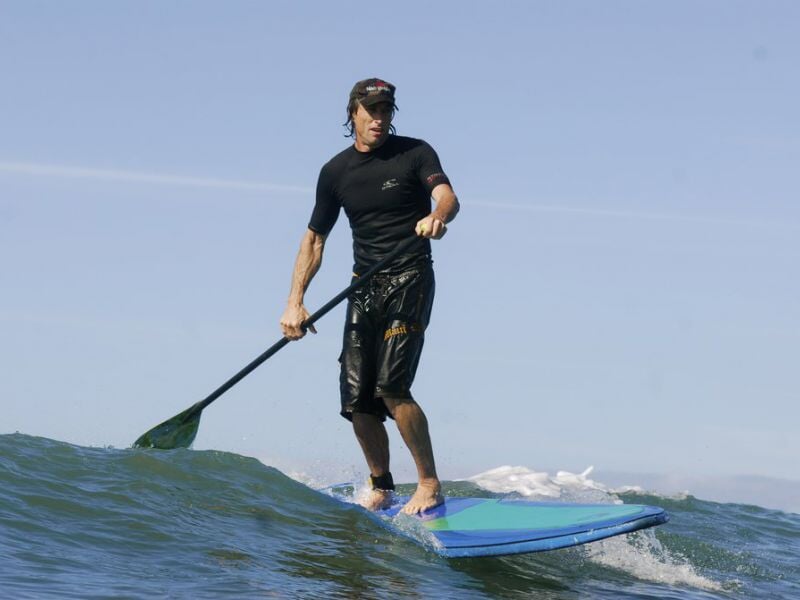 Homme en combinaison noire debout sur une planche de paddle surf, pagayant sur une vague.