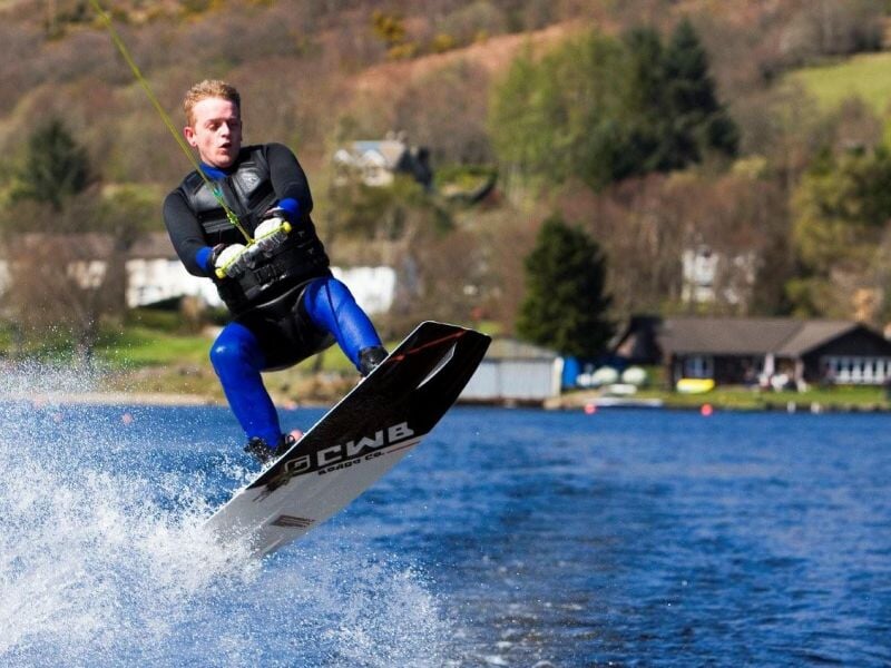Homme en combinaison bleue effectuant un saut spectaculaire en wakeboard sur un lac.