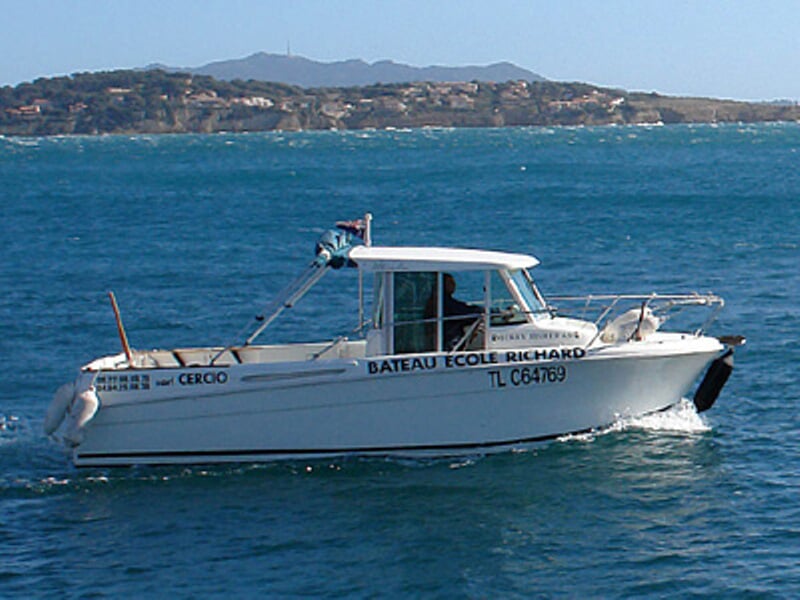 Bateau école Richard naviguant sur une mer calme avec un paysage côtier en arrière-plan sous un ciel clair.