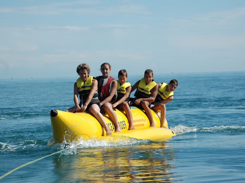 Cinq personnes portant des gilets de sauvetage jaunes et rouges s'amusent sur un bateau banane jaune sur l'eau bleue.