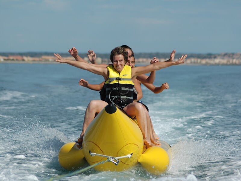 Trois personnes souriantes portant des gilets de sauvetage jaunes font du bateau banane sur une mer calme sous un ciel bleu.