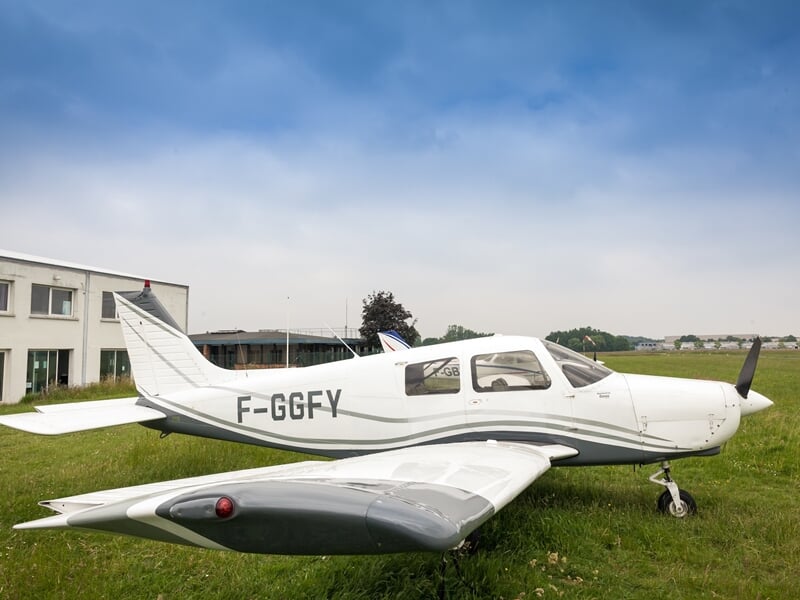 Petit avion blanc immatriculé F-GGFY stationné sur l'herbe devant un bâtiment sous un ciel partiellement nuageux.