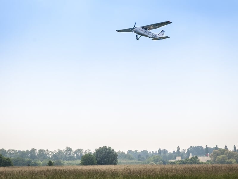 Avion léger en vol au-dessus d'un champ avec des arbres et des bâtiments à l'horizon sous un ciel clair.