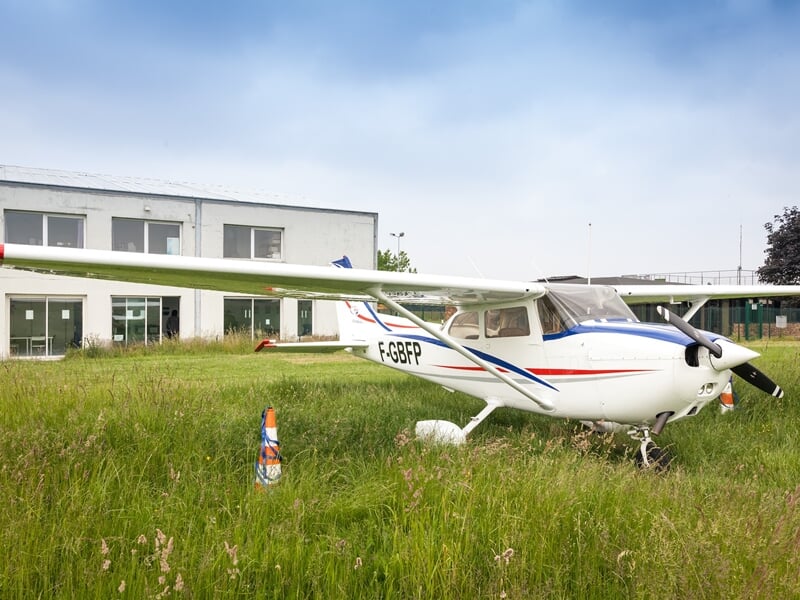 Avion blanc avec des bandes bleues et rouges garé sur une pelouse verte devant un bâtiment industriel.