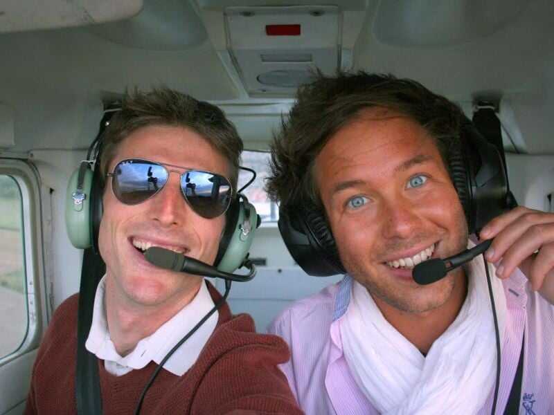 Deux hommes souriants avec casques de pilote dans un avion, l'un porte des lunettes de soleil réfléchissantes.