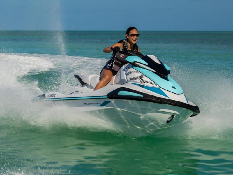 Femme souriante en gilet de sauvetage pilotant un jet ski Yamaha sur une mer turquoise, générant une traînée d'eau derrière elle. Loisirs nautiques, aventure en plein air, et plaisir en solo sous un c