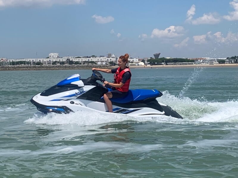 Femme en gilet de sauvetage rouge pilotant un jet-ski Yamaha bleu et blanc, créant des éclaboussures sur une mer calme. En arrière-plan, une plage avec des immeubles sous un ciel partiellement nuageux