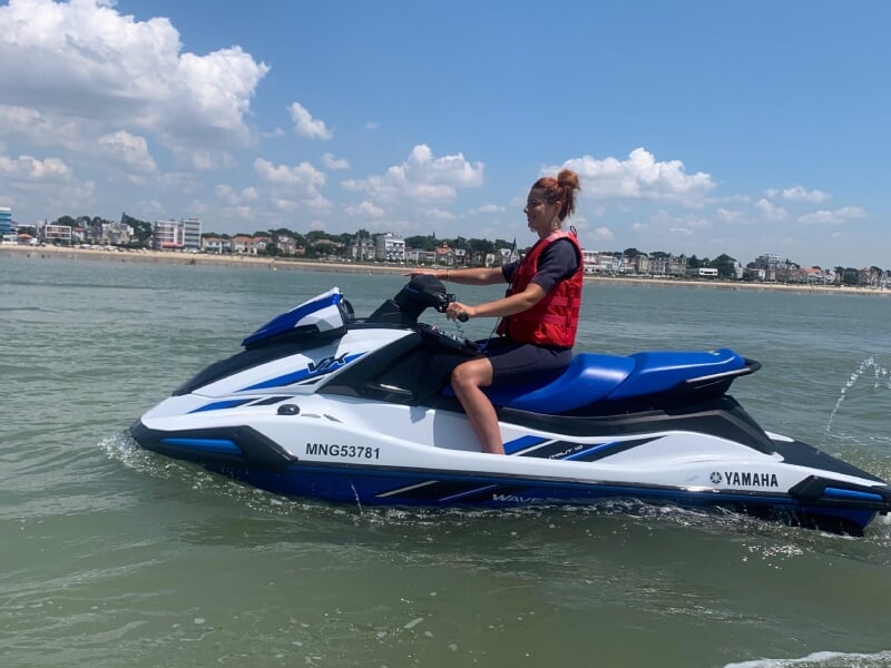 Femme en gilet de sauvetage rouge conduisant un jet-ski Yamaha bleu et blanc sur une mer calme. En arrière-plan, une plage avec des immeubles sous un ciel partiellement nuageux. Activité nautique esti