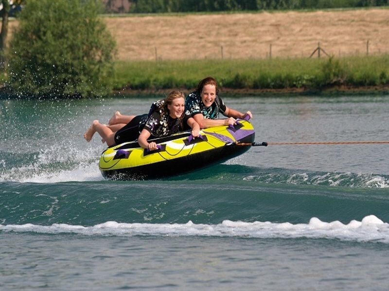 Deux jeunes femmes s'amusent sur une bouée tractée jaune et noire sur un lac, éclaboussures d'eau autour.