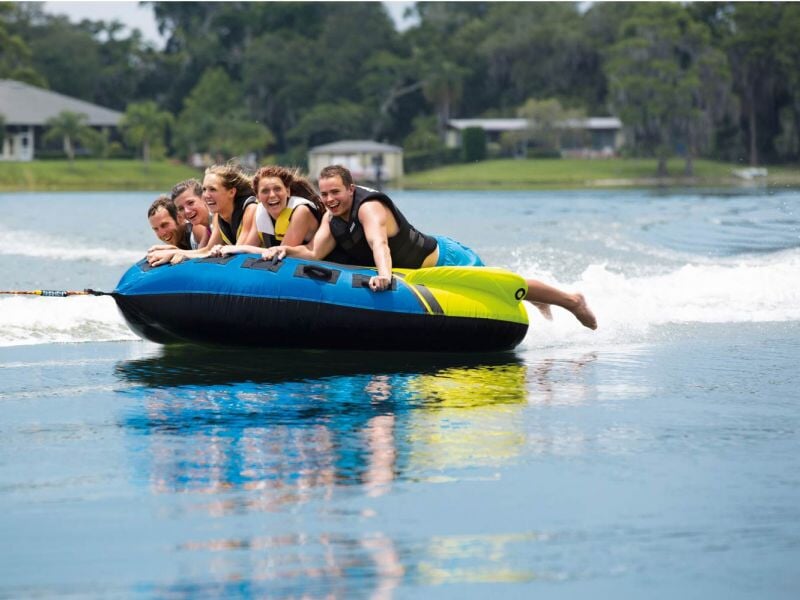 Groupe de cinq personnes souriantes sur une grande bouée bleue et jaune tractée sur un lac calme, ambiance joyeuse.