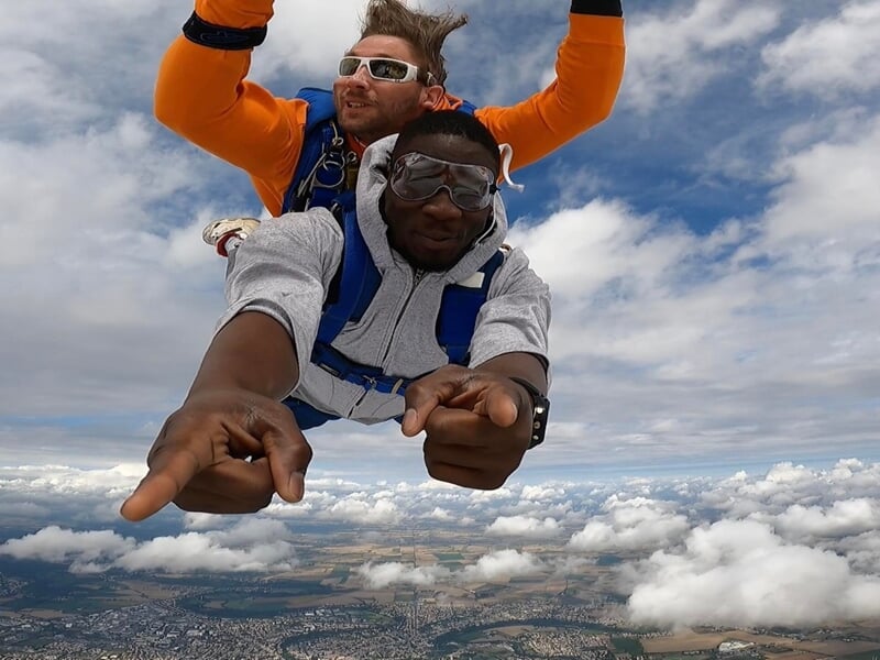 Deux parachutistes en tandem en chute libre au-dessus d'une ville, avec un ciel nuageux et lumineux.