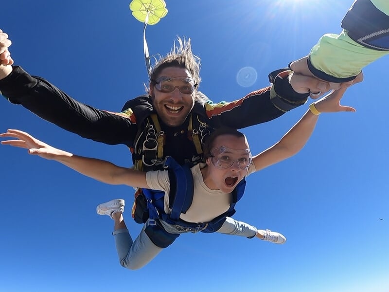 Parachutisme tandem sous le soleil, parachute ouvert en arrière-plan, ambiance de liberté.