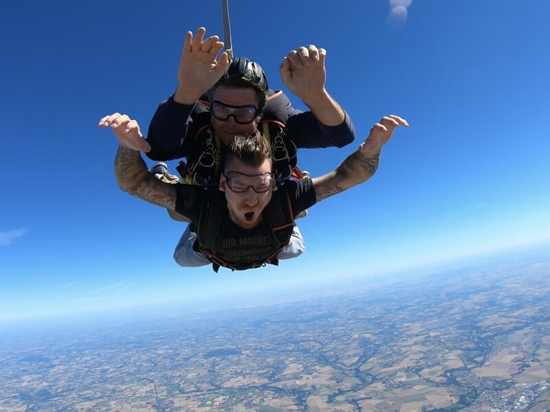 Chute libre tandem sous un ciel bleu, expressions d’euphorie, vue panoramique sur la campagne.