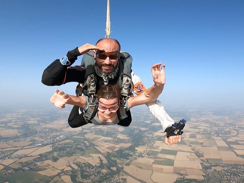 Saut tandem au-dessus d’une campagne, instructeur saluant et passagère bras écartés, ciel clair.