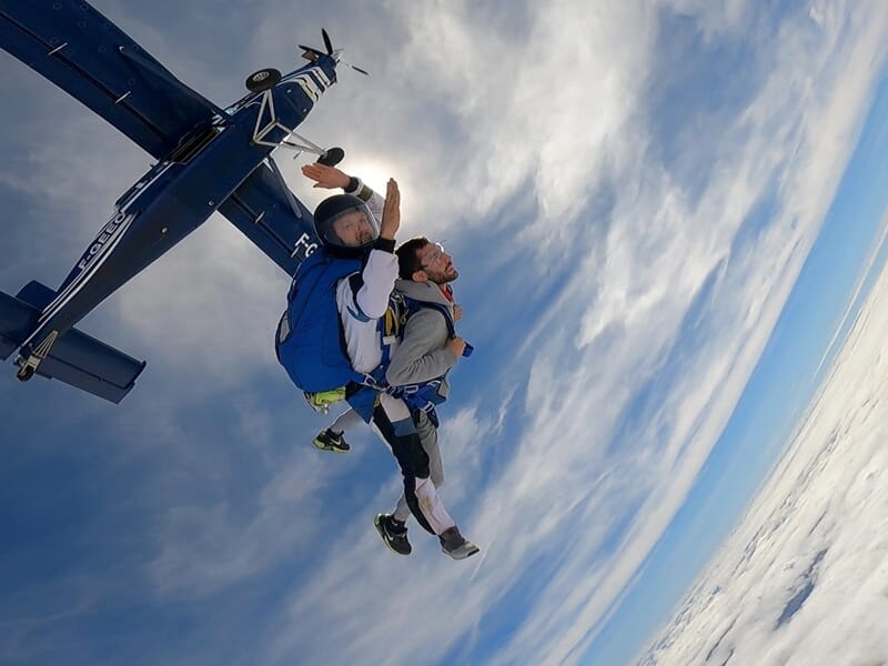 Chute libre en tandem près d’un avion bleu, ciel dégagé avec nuages en altitude. Sensation d’adrénaline.