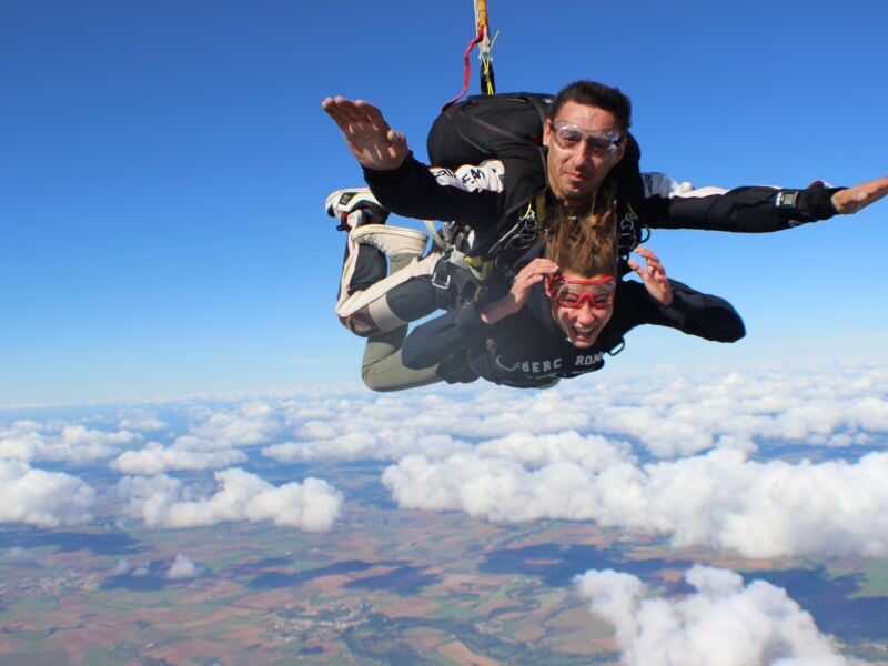 Deux personnes en saut en parachute en tandem, volant au-dessus des nuages avec un paysage terrestre en arrière-plan.
