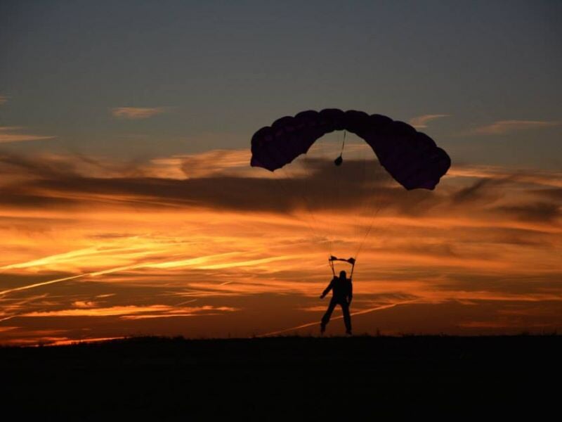 Parachutiste silhouetté au sol avec un parachute déployé, au coucher du soleil aux couleurs orangées.