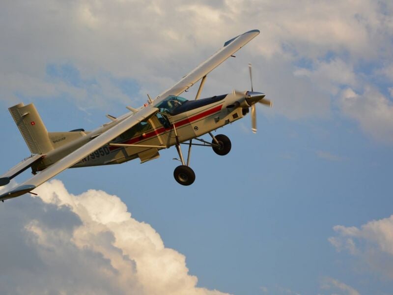 Petit avion blanc avec des bandes colorées volant dans un ciel partiellement nuageux, prêt pour un saut.