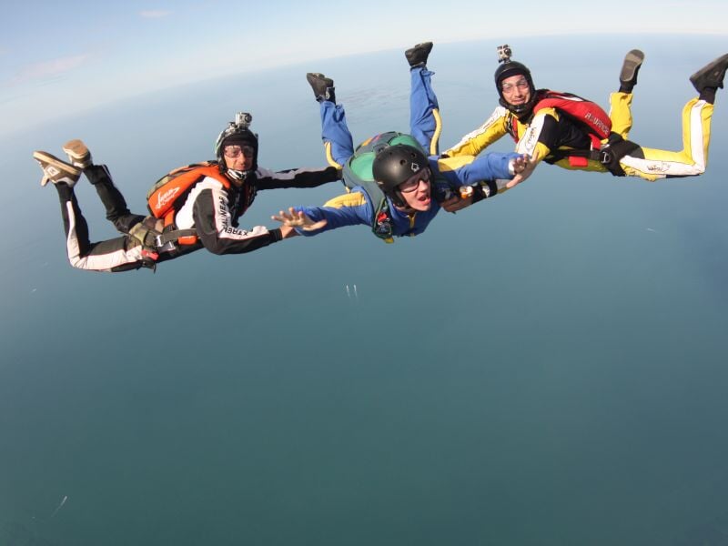 Trois parachutistes en formation en chute libre, tenant leurs mains dans un ciel bleu au-dessus de la mer.