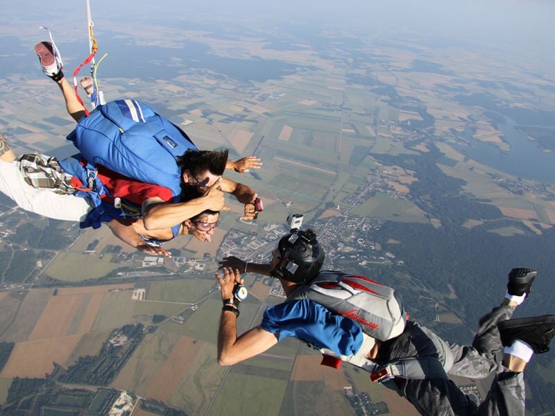 Trois parachutistes en chute libre, deux en bleu et un en rouge, volant au-dessus d'un paysage agricole.