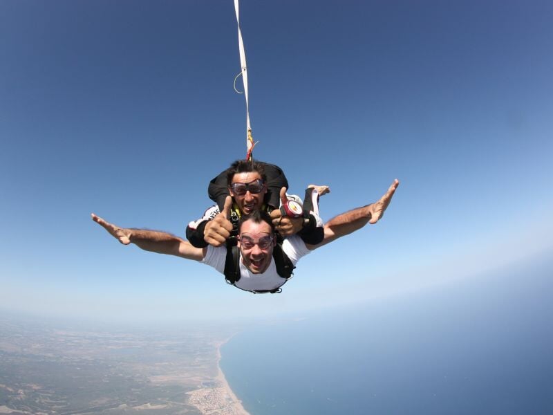 Deux parachutistes en tandem souriants en chute libre, avec un paysage côtier visible en dessous.