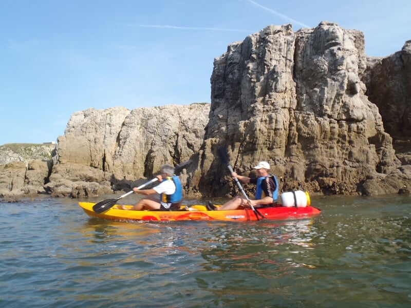 Deux personnes pagayant dans un kayak rouge et jaune près de falaises rocheuses au bord de la mer.