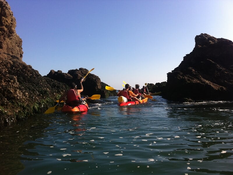 Groupe de personnes pagayant dans des kayaks rouges entre des rochers sombres sur une rivière étroite.