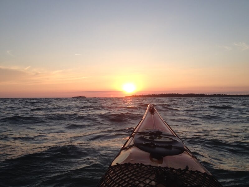 Vue depuis un kayak sur l'eau avec un coucher de soleil à l'horizon sur une mer calme.