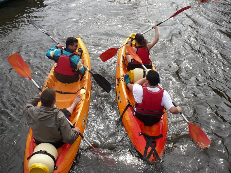 Quatre personnes pagayant dans deux kayaks jaunes et rouges sur une rivière entourée d'arbres verts.