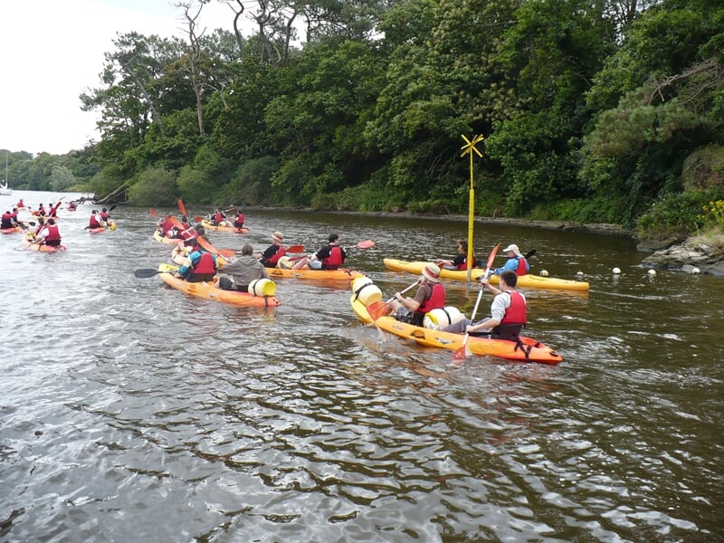Groupe de plusieurs kayaks jaunes et rouges pagayant sur une rivière calme avec des arbres en arrière-plan.