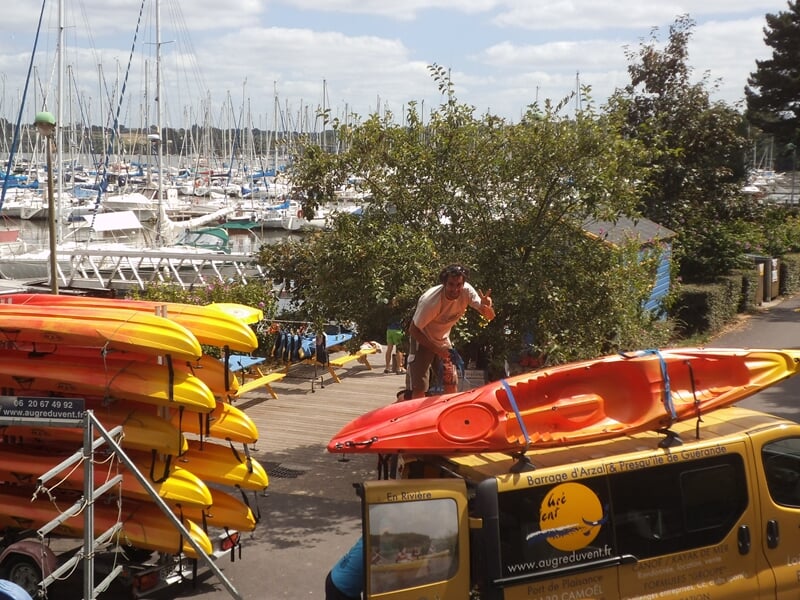 Homme pagayant dans un kayak rouge et jaune sur une rivière entourée d'arbres verts et de buissons.