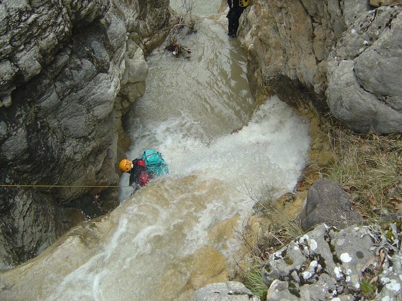 Personne équipée d'un casque et d'un sac à dos descend une cascade rocheuse en canyoning dans un environnement naturel.