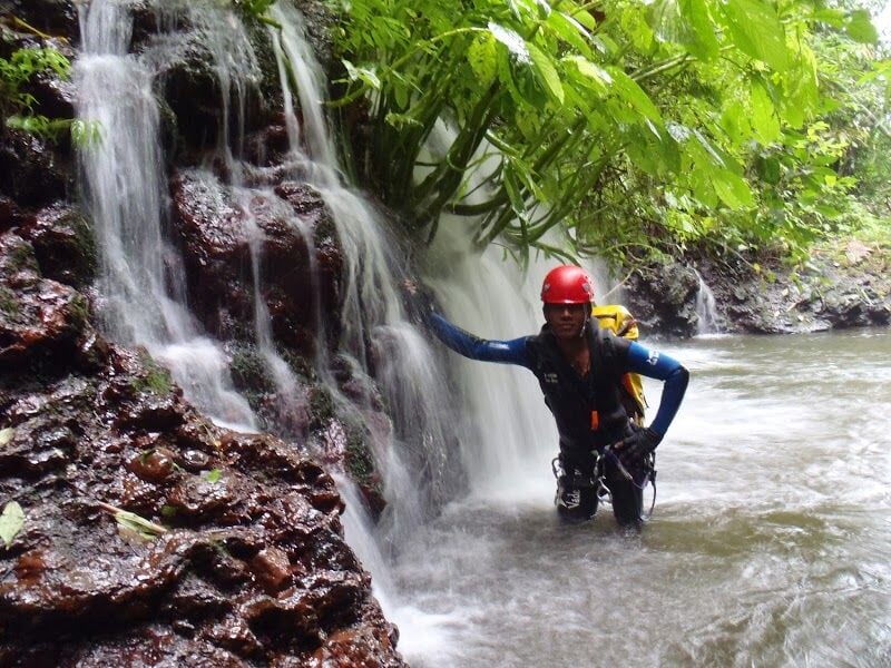 Personne en casque rouge et combinaison noire explore une cascade d'eau dans un cadre naturel verdoyant.