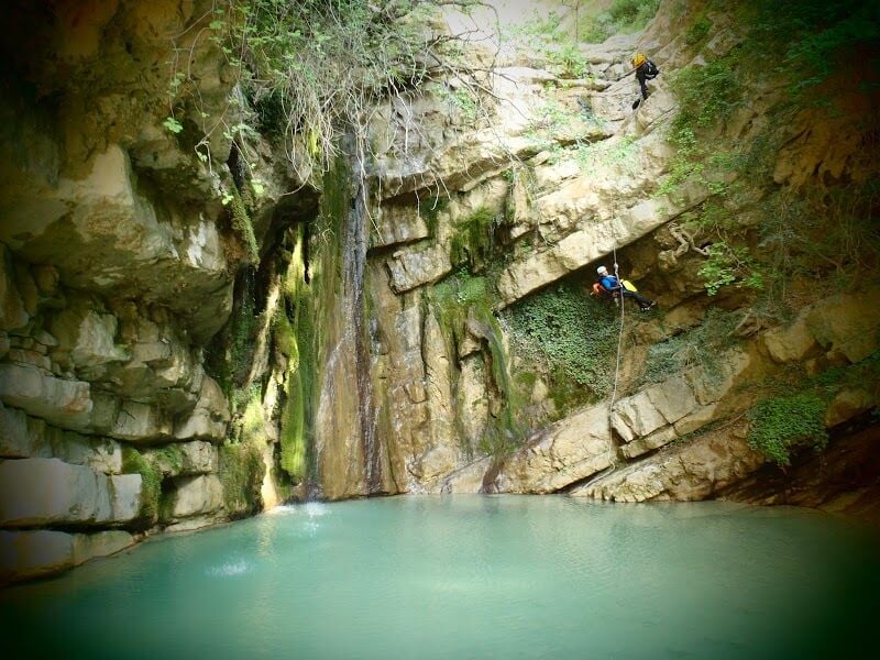 Deux personnes en équipement de canyoning descendent en rappel une falaise au-dessus d'un bassin d'eau turquoise.