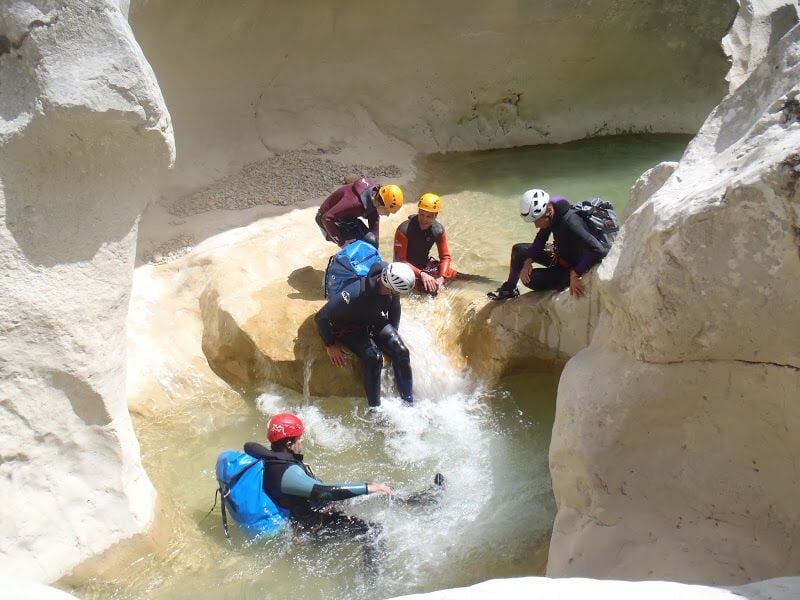 Groupe de cinq personnes en combinaison et casque se reposent dans un bassin d'eau claire entouré de rochers.