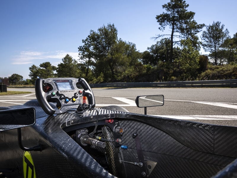 Intérieur d'une Formule Renault avec volant et tableau de bord sur circuit entouré d'arbres.