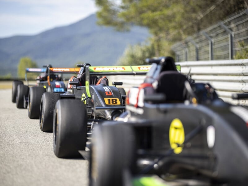 Alignement de plusieurs Formule Renault noires sur piste avec pilotes casqués et barrières métalliques.