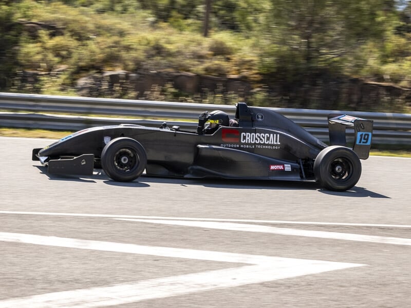 Formule Renault noire numéro 19 en virage sur piste avec barrières de sécurité et arbres autour.