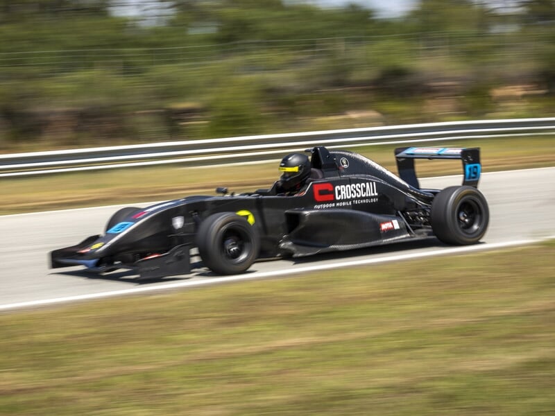 Formule Renault noire numéro 18 en virage rapide sur piste avec pilote casqué et herbe en bordure.