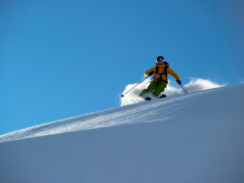 Skieur en tenue jaune et verte dévalant une pente enneigée sous un ciel bleu clair sans nuages.