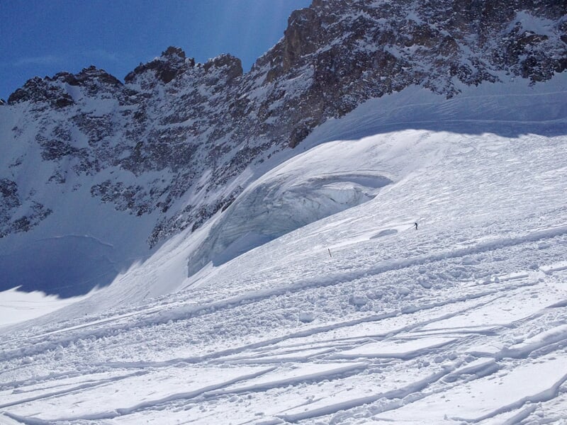 Pente de montagne enneigée avec traces de ski et un glacier visible sous un ciel bleu profond.
